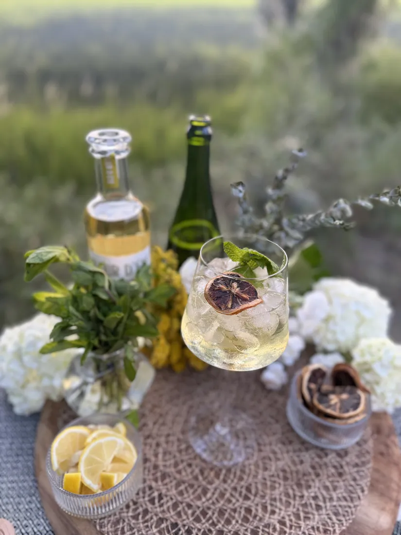 A sparkling elderflower spritz with fresh mint and a dried citrus wheel, styled on a wooden board with hydrangeas, lemons and bottles in a sunlit field